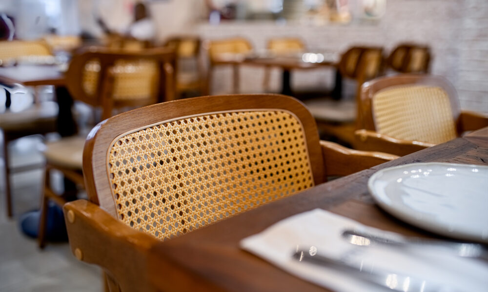 Uma foto em primeiro plano e com foco seletivo mostra uma cadeira de madeira com encosto de palha trançada clara, posicionada junto a uma mesa de madeira escura em um restaurante. Sobre a mesa, veem-se parcialmente um prato branco e talheres embrulhados em um guardanapo. O fundo da imagem está desfocado, revelando outras mesas e cadeiras semelhantes espalhadas pelo salão