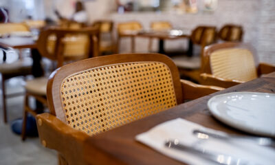 Uma foto em primeiro plano e com foco seletivo mostra uma cadeira de madeira com encosto de palha trançada clara, posicionada junto a uma mesa de madeira escura em um restaurante. Sobre a mesa, veem-se parcialmente um prato branco e talheres embrulhados em um guardanapo. O fundo da imagem está desfocado, revelando outras mesas e cadeiras semelhantes espalhadas pelo salão