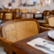 Uma foto em primeiro plano e com foco seletivo mostra uma cadeira de madeira com encosto de palha trançada clara, posicionada junto a uma mesa de madeira escura em um restaurante. Sobre a mesa, veem-se parcialmente um prato branco e talheres embrulhados em um guardanapo. O fundo da imagem está desfocado, revelando outras mesas e cadeiras semelhantes espalhadas pelo salão