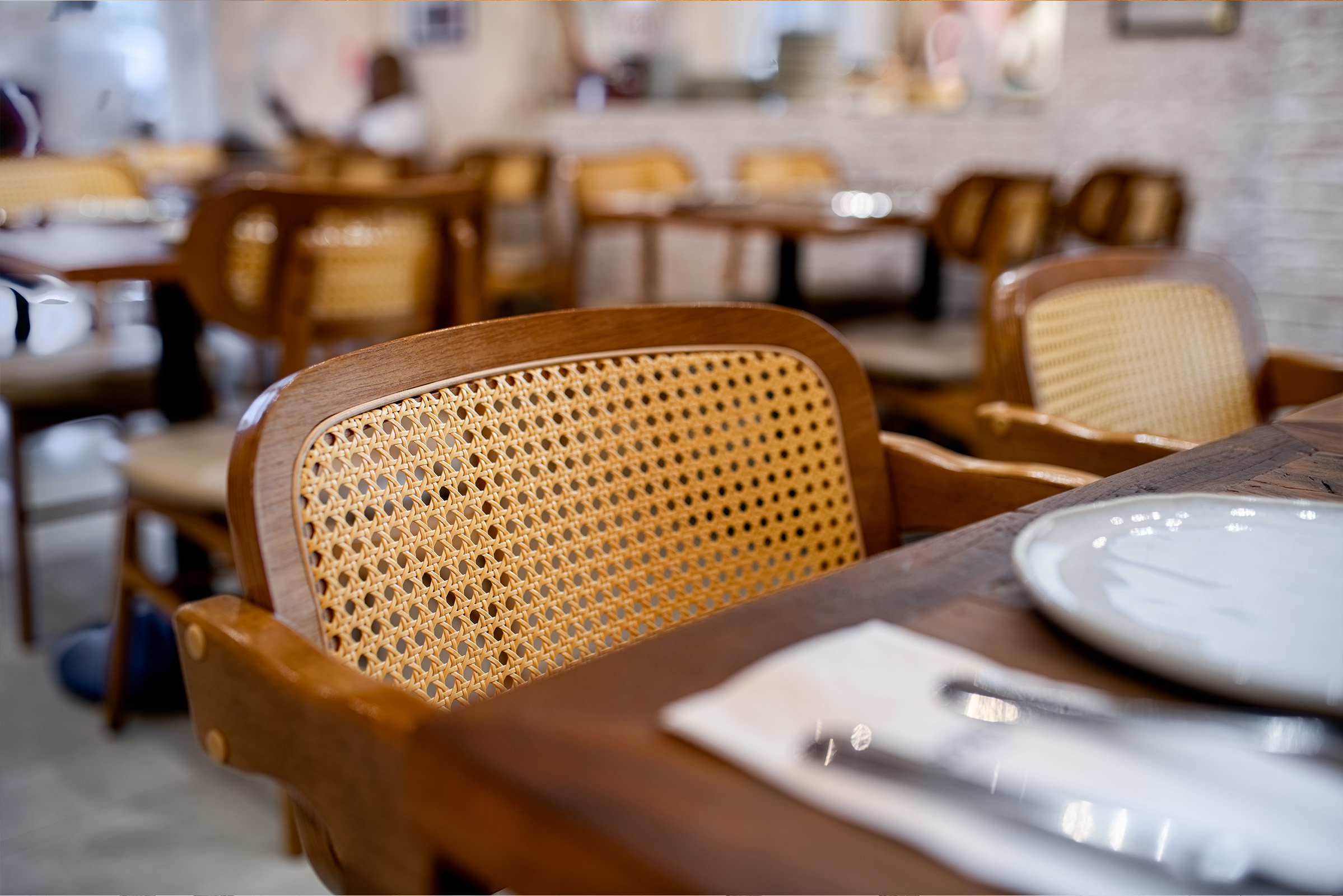 Uma foto em primeiro plano e com foco seletivo mostra uma cadeira de madeira com encosto de palha trançada clara, posicionada junto a uma mesa de madeira escura em um restaurante. Sobre a mesa, veem-se parcialmente um prato branco e talheres embrulhados em um guardanapo. O fundo da imagem está desfocado, revelando outras mesas e cadeiras semelhantes espalhadas pelo salão