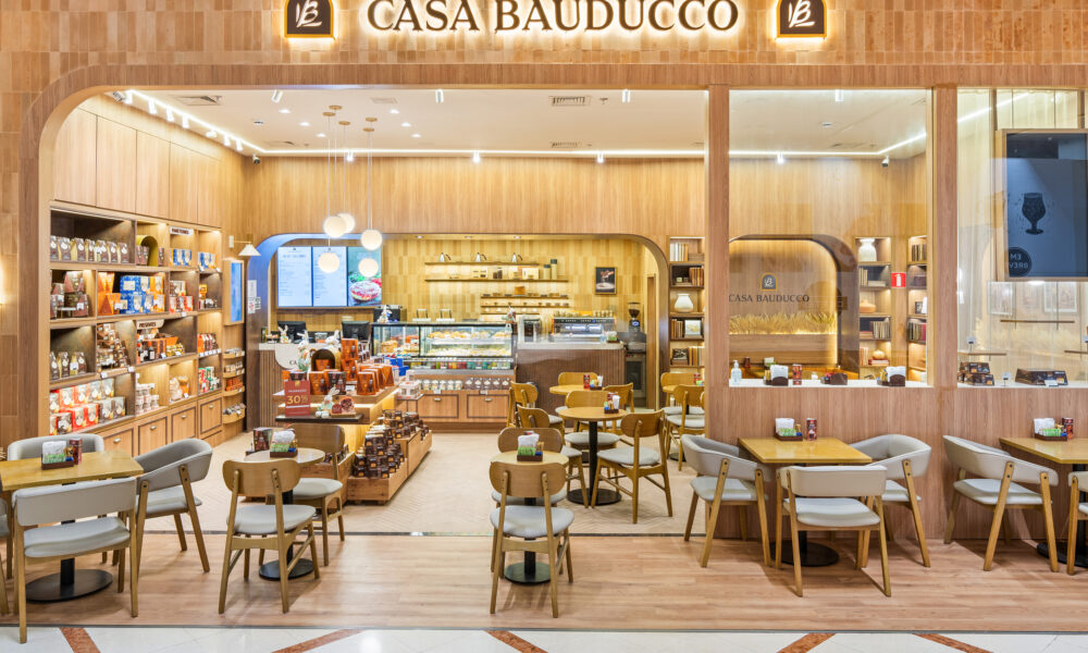 Fachada da cafeteria Casa Bauducco localizada em um shopping. A loja possui acabamento em tons de madeira clara, iluminação aconchegante e mesas com cadeiras estofadas dispostas na área externa e interna. Vasos horizontais com ramos de trigo decoram as laterais da entrada.