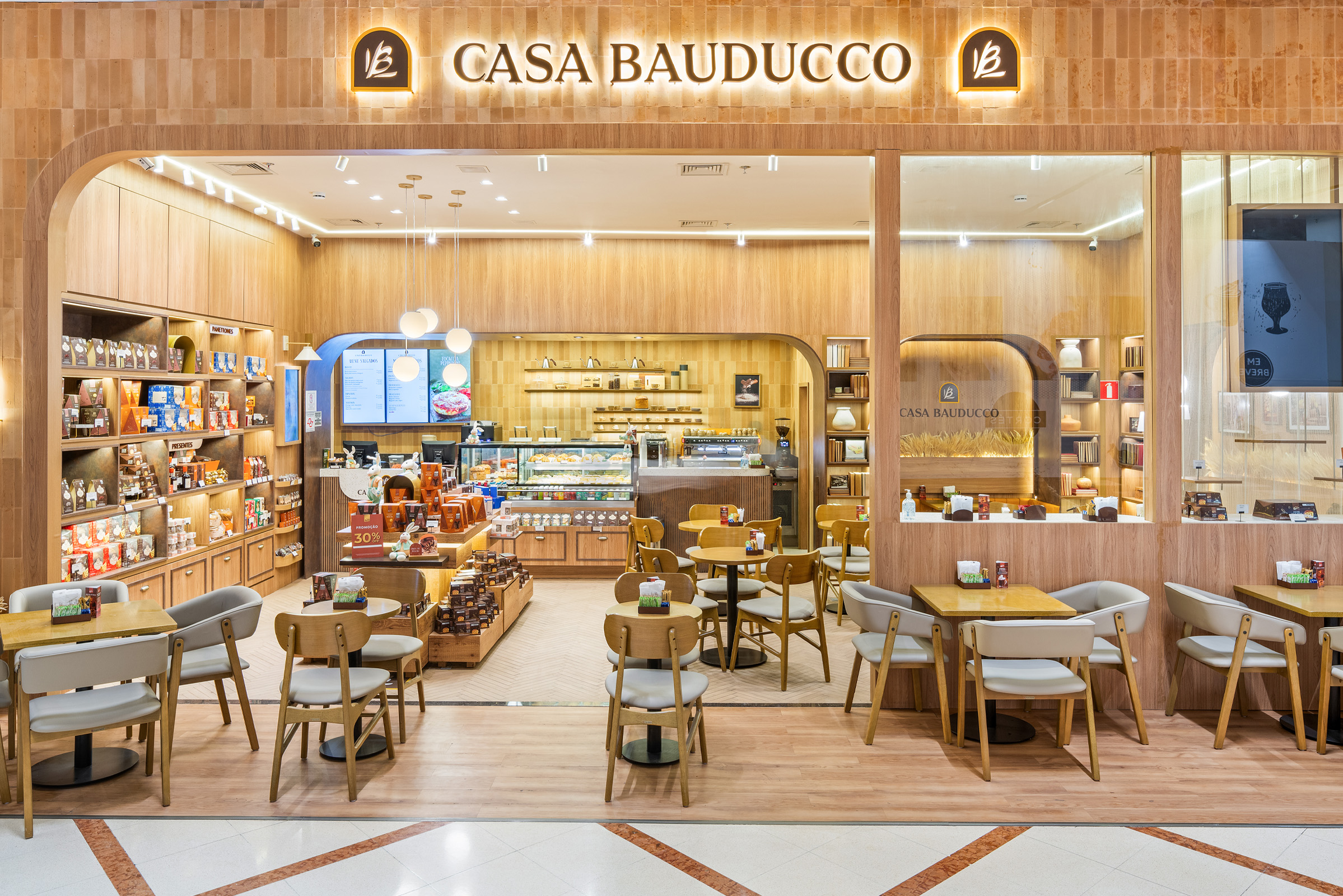 Fachada da cafeteria Casa Bauducco localizada em um shopping. A loja possui acabamento em tons de madeira clara, iluminação aconchegante e mesas com cadeiras estofadas dispostas na área externa e interna. Vasos horizontais com ramos de trigo decoram as laterais da entrada.