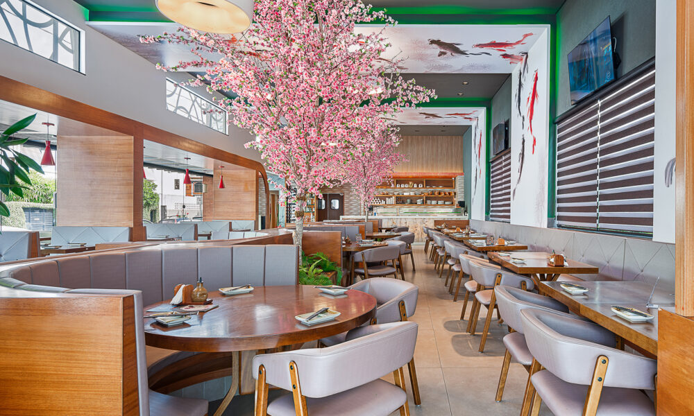 Uma imagem panorâmica do interior de um restaurante contemporâneo com decoração de inspiração asiática. O salão apresenta mesas redondas e retangulares de madeira acompanhadas por cadeiras e sofás estofados em tons claros de cinza. Destacam-se no ambiente árvores ornamentais com flores rosa artificiais, lembrando cerejeiras, além de paredes brancas decoradas com pinturas de carpas e detalhes em madeira vazada.