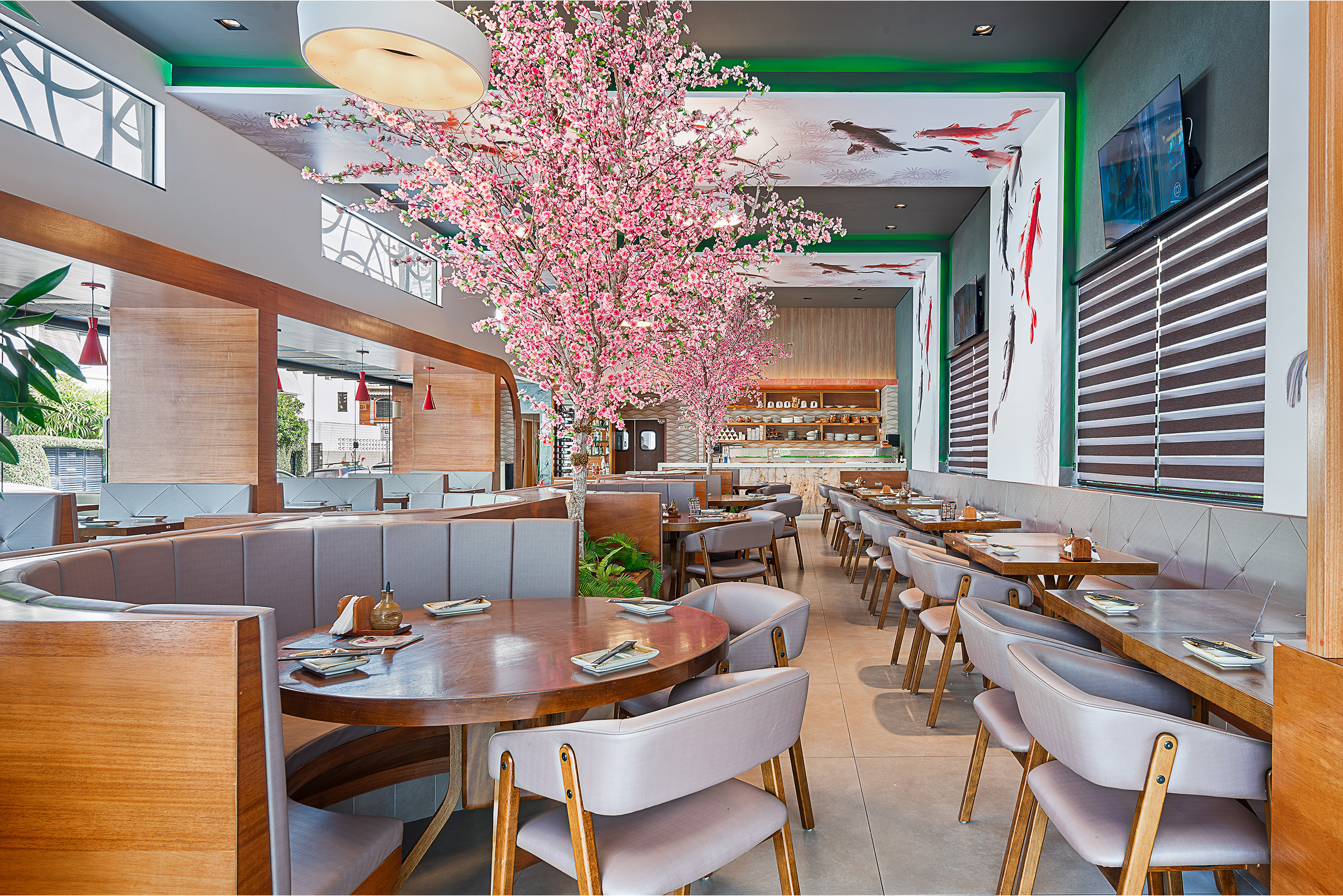 Uma imagem panorâmica do interior de um restaurante contemporâneo com decoração de inspiração asiática. O salão apresenta mesas redondas e retangulares de madeira acompanhadas por cadeiras e sofás estofados em tons claros de cinza. Destacam-se no ambiente árvores ornamentais com flores rosa artificiais, lembrando cerejeiras, além de paredes brancas decoradas com pinturas de carpas e detalhes em madeira vazada.