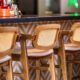 Fotografia em foco em uma fileira de banquetas altas de madeira com encosto de palha trançada, alinhadas ao longo de um balcão de bar. O balcão possui tampo escuro e frente decorada com azulejos geométricos em zigue-zague. Ao fundo, o ambiente do bar aparece levemente desfocado.