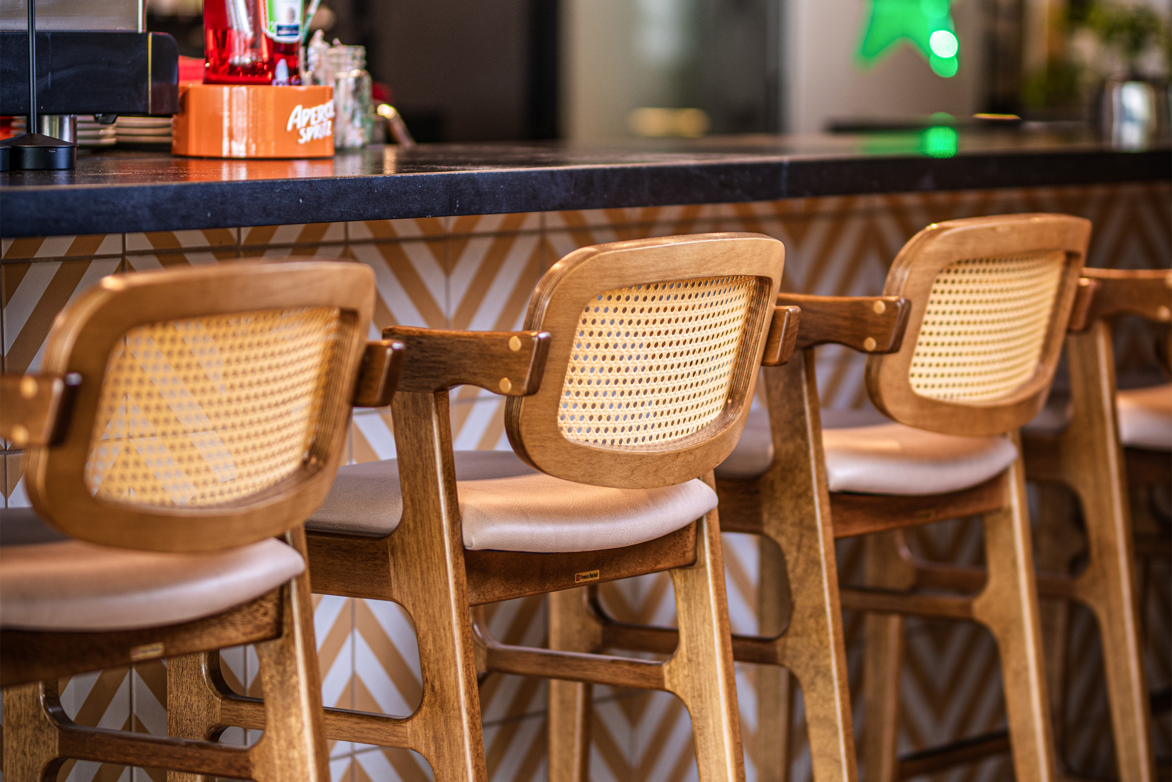 Fotografia em foco em uma fileira de banquetas altas de madeira com encosto de palha trançada, alinhadas ao longo de um balcão de bar. O balcão possui tampo escuro e frente decorada com azulejos geométricos em zigue-zague. Ao fundo, o ambiente do bar aparece levemente desfocado.