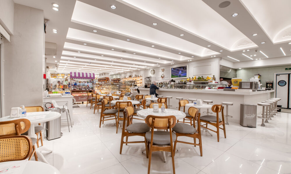 Interior de um restaurante amplo, luminoso e predominantemente branco. O salão é preenchido por várias mesas brancas quadradas com cadeiras de madeira de tom médio e estofamento cinza. Grandes colunas redondas brancas sustentam o teto, que possui sancas de gesso com iluminação embutida em linhas modernas e circulares. 