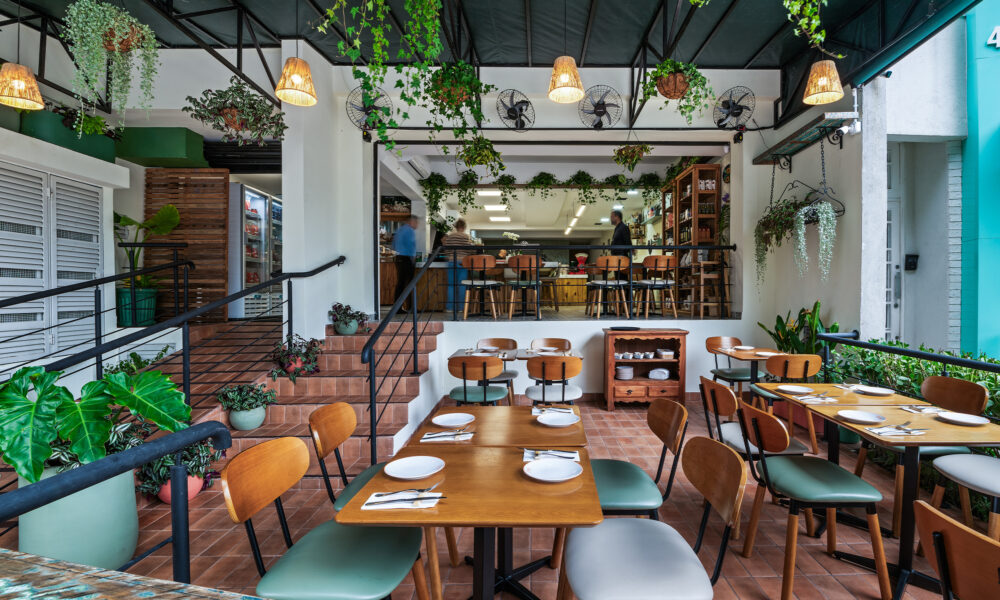 Área externa coberta de um restaurante acolhedor, decorada com muitas plantas pendentes e vasos verdes. O espaço tem piso de lajota cerâmica terracota e mesas de madeira com pratos e talheres arrumados. As cadeiras misturam encostos de madeira com assentos estofados em tons de verde-escuro e cinza-claro. À esquerda, uma rampa e uma pequena escada com corrimão preto levam ao salão interno elevado, onde há um balcão com banquetas e funcionários trabalhando ao fundo. Lustres de palha trançada iluminam o ambiente sob uma cobertura escura.