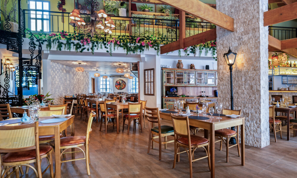 Interior de um restaurante amplo com decoração rústica e acolhedora. No salão principal, há diversas mesas de madeira com cadeiras de encosto de palha dispostas sobre o piso amadeirado. Grandes pilares revestidos de pedras claras sustentam robustas vigas de madeira expostas no teto. Ao fundo, à direita, destaca-se um balcão de bar também revestido de pedra com banquetas altas de madeira. A iluminação quente e suave vem de lustres clássicos e postes de luz internos de estilo retrô.