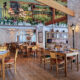 Interior de um restaurante amplo com decoração rústica e acolhedora. No salão principal, há diversas mesas de madeira com cadeiras de encosto de palha dispostas sobre o piso amadeirado. Grandes pilares revestidos de pedras claras sustentam robustas vigas de madeira expostas no teto. Ao fundo, à direita, destaca-se um balcão de bar também revestido de pedra com banquetas altas de madeira. A iluminação quente e suave vem de lustres clássicos e postes de luz internos de estilo retrô.