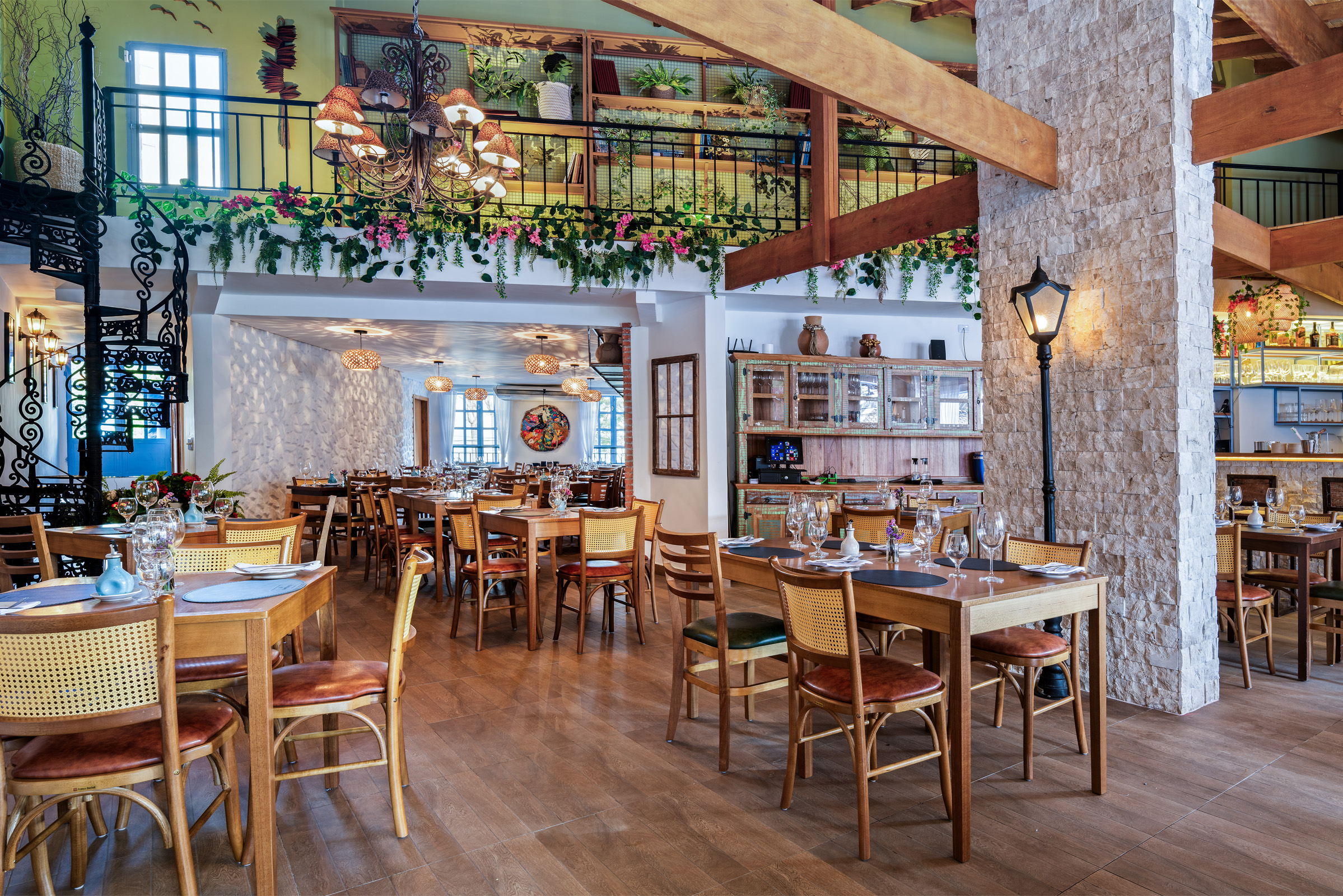 Interior de um restaurante amplo com decoração rústica e acolhedora. No salão principal, há diversas mesas de madeira com cadeiras de encosto de palha dispostas sobre o piso amadeirado. Grandes pilares revestidos de pedras claras sustentam robustas vigas de madeira expostas no teto. Ao fundo, à direita, destaca-se um balcão de bar também revestido de pedra com banquetas altas de madeira. A iluminação quente e suave vem de lustres clássicos e postes de luz internos de estilo retrô.