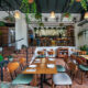 Área externa coberta de um restaurante acolhedor, decorada com muitas plantas pendentes e vasos verdes. O espaço tem piso de lajota cerâmica terracota e mesas de madeira com pratos e talheres arrumados. As cadeiras misturam encostos de madeira com assentos estofados em tons de verde-escuro e cinza-claro. À esquerda, uma rampa e uma pequena escada com corrimão preto levam ao salão interno elevado, onde há um balcão com banquetas e funcionários trabalhando ao fundo. Lustres de palha trançada iluminam o ambiente sob uma cobertura escura.