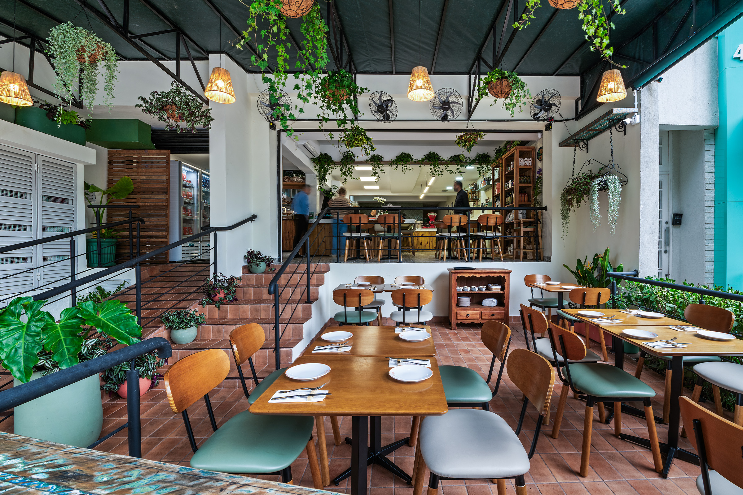 Área externa coberta de um restaurante acolhedor, decorada com muitas plantas pendentes e vasos verdes. O espaço tem piso de lajota cerâmica terracota e mesas de madeira com pratos e talheres arrumados. As cadeiras misturam encostos de madeira com assentos estofados em tons de verde-escuro e cinza-claro. À esquerda, uma rampa e uma pequena escada com corrimão preto levam ao salão interno elevado, onde há um balcão com banquetas e funcionários trabalhando ao fundo. Lustres de palha trançada iluminam o ambiente sob uma cobertura escura.