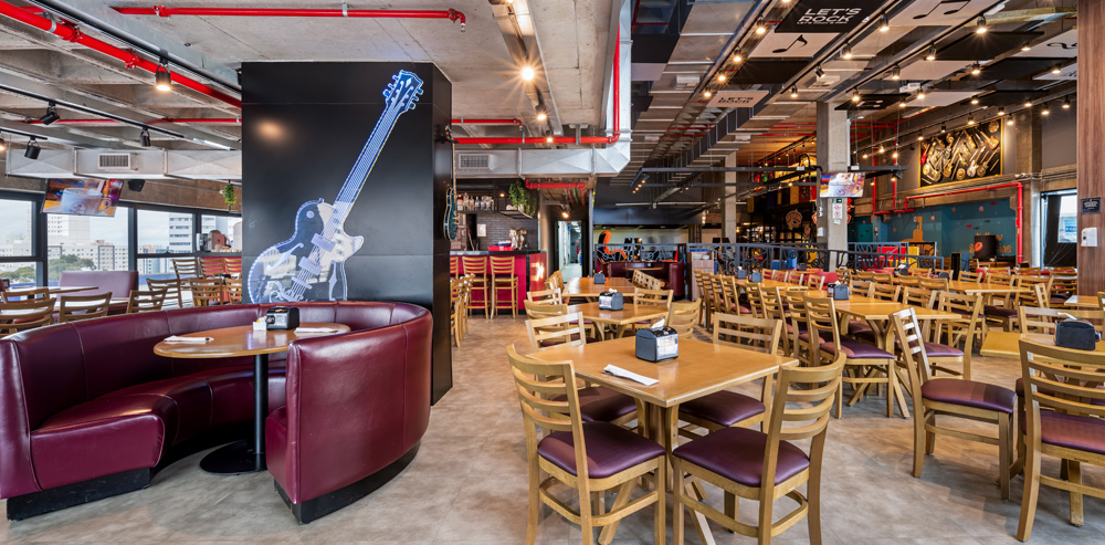Amplo salão de um restaurante temático de rock com estilo industrial. No primeiro plano, à esquerda, há uma mesa redonda de madeira cercada por um sofá curvo estofado em couro bordô, posicionado em frente a um grande pilar preto decorado com a ilustração de uma guitarra elétrica. O restante do salão é preenchido por diversas mesas quadradas de madeira com cadeiras de assento roxo. O teto apresenta vigas de concreto expostas, tubulações vermelhas de incêndio, trilhos de iluminação preta e, ao fundo, painéis decorativos com notas musicais e a frase "Let's Rock". Ao fundo, à esquerda, há um balcão de bar iluminado em vermelho e grandes janelas com vista para a cidade.
