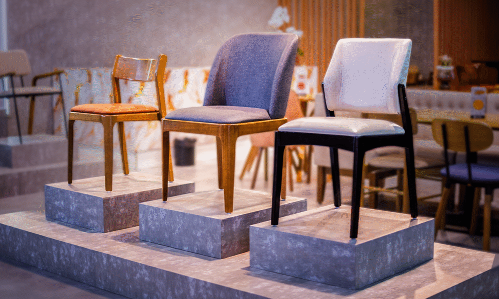 Exposição de cadeiras de design moderno dispostas sobre três pedestais cinzas de alturas diferentes. No pedestal da direita, destaca-se uma cadeira com estofamento branco e pés de madeira pintada de preto. No pedestal central, há uma cadeira estofada em tecido cinza-azulável com pés de madeira em tom natural médio. No pedestal da esquerda, observa-se uma cadeira simples de madeira clara com assento estofado em tom caramelo.