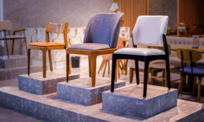 Exposição de cadeiras de design moderno dispostas sobre três pedestais cinzas de alturas diferentes. No pedestal da direita, destaca-se uma cadeira com estofamento branco e pés de madeira pintada de preto. No pedestal central, há uma cadeira estofada em tecido cinza-azulável com pés de madeira em tom natural médio. No pedestal da esquerda, observa-se uma cadeira simples de madeira clara com assento estofado em tom caramelo.