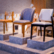 Exposição de cadeiras de design moderno dispostas sobre três pedestais cinzas de alturas diferentes. No pedestal da direita, destaca-se uma cadeira com estofamento branco e pés de madeira pintada de preto. No pedestal central, há uma cadeira estofada em tecido cinza-azulável com pés de madeira em tom natural médio. No pedestal da esquerda, observa-se uma cadeira simples de madeira clara com assento estofado em tom caramelo.