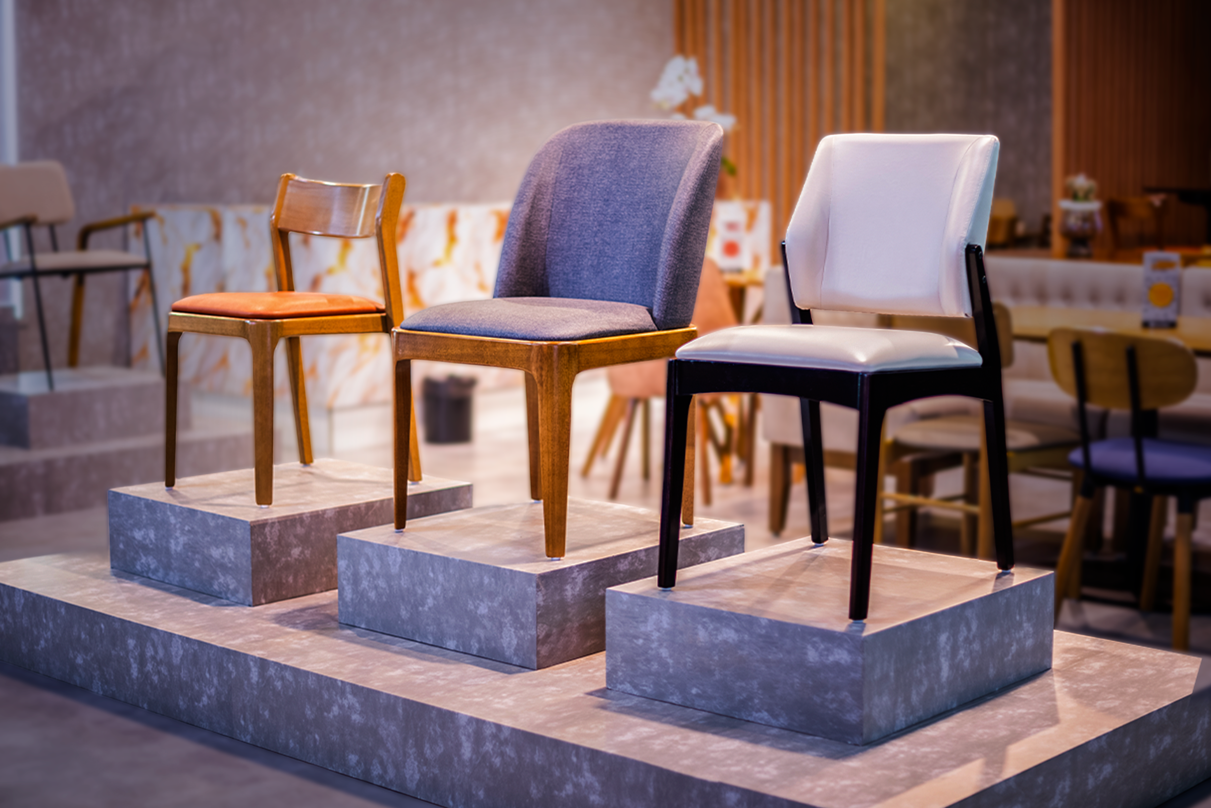 Exposição de cadeiras de design moderno dispostas sobre três pedestais cinzas de alturas diferentes. No pedestal da direita, destaca-se uma cadeira com estofamento branco e pés de madeira pintada de preto. No pedestal central, há uma cadeira estofada em tecido cinza-azulável com pés de madeira em tom natural médio. No pedestal da esquerda, observa-se uma cadeira simples de madeira clara com assento estofado em tom caramelo.