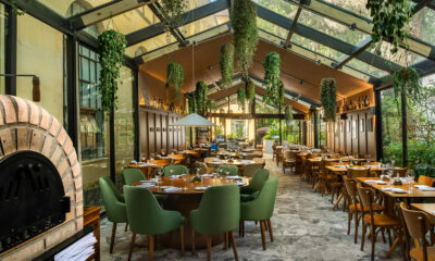 Restaurante amplo com teto de vidro, muita iluminação natural e plantas suspensas. O salão conta com mesas de madeira, cadeiras verdes e cadeiras em madeira, distribuídas ao longo do espaço. À esquerda, há um forno de tijolos em destaque, enquanto o fundo mostra vegetação e elementos em madeira que reforçam a atmosfera acolhedora do ambiente.