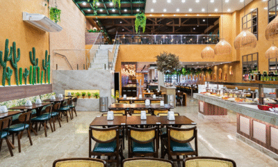 Vista geral do salão de um restaurante de buffet amplo e bem iluminado com decoração rústica e moderna. À direita, estende-se um longo balcão de buffet self-service com tampo de pedra clara, oferecendo diversas opções de pratos quentes e saladas em travessas coloridas; acima dele, há uma decoração inusitada com uma bicicleta verde suspensa. No centro e à esquerda, organizam-se várias mesas de madeira com cadeiras de encosto de palha e assentos escuros.