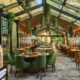 Restaurante amplo com teto de vidro, muita iluminação natural e plantas suspensas. O salão conta com mesas de madeira, cadeiras verdes e cadeiras em madeira, distribuídas ao longo do espaço. À esquerda, há um forno de tijolos em destaque, enquanto o fundo mostra vegetação e elementos em madeira que reforçam a atmosfera acolhedora do ambiente.