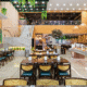 Vista geral do salão de um restaurante de buffet amplo e bem iluminado com decoração rústica e moderna. À direita, estende-se um longo balcão de buffet self-service com tampo de pedra clara, oferecendo diversas opções de pratos quentes e saladas em travessas coloridas; acima dele, há uma decoração inusitada com uma bicicleta verde suspensa. No centro e à esquerda, organizam-se várias mesas de madeira com cadeiras de encosto de palha e assentos escuros.