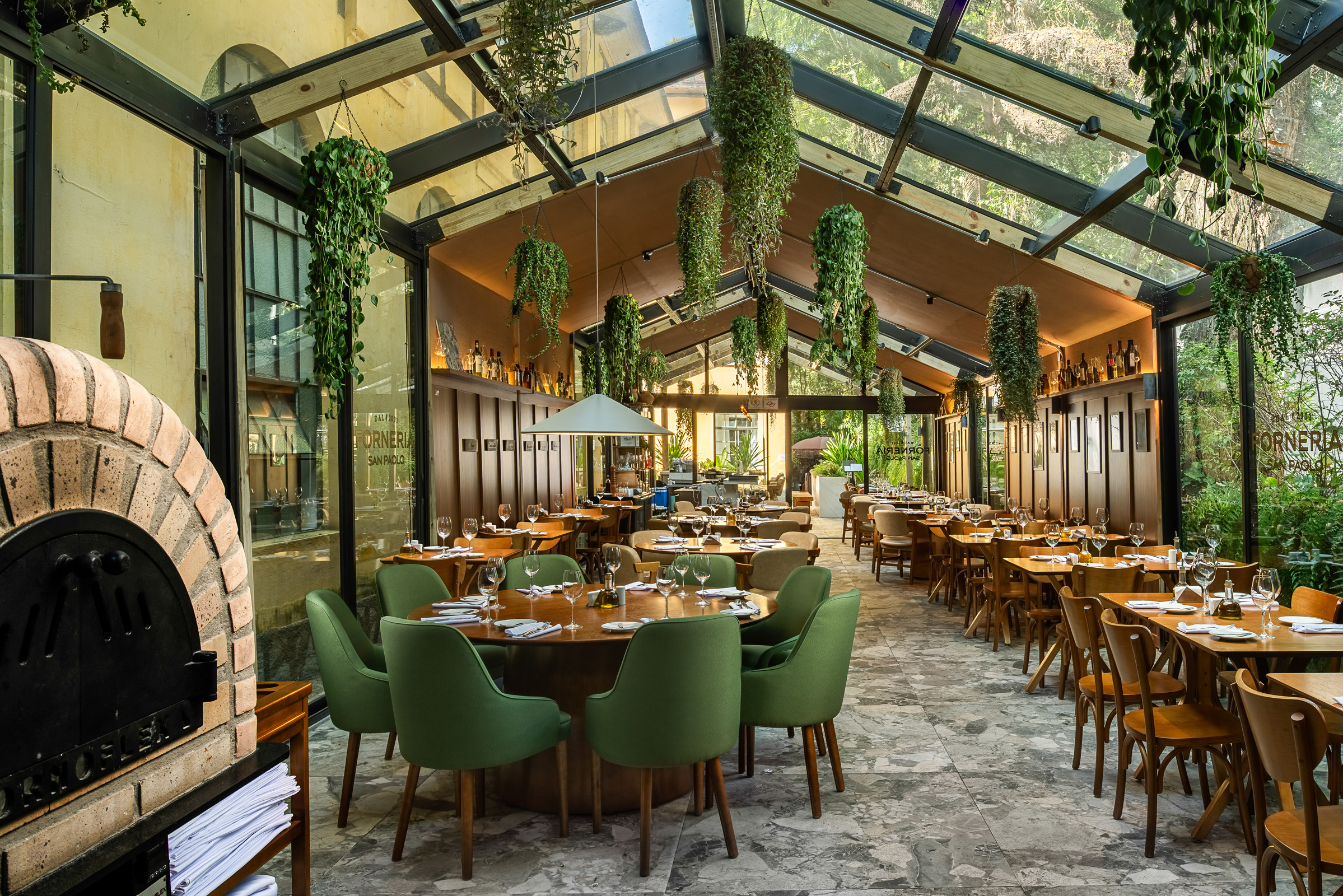 Restaurante amplo com teto de vidro, muita iluminação natural e plantas suspensas. O salão conta com mesas de madeira, cadeiras verdes e cadeiras em madeira, distribuídas ao longo do espaço. À esquerda, há um forno de tijolos em destaque, enquanto o fundo mostra vegetação e elementos em madeira que reforçam a atmosfera acolhedora do ambiente.