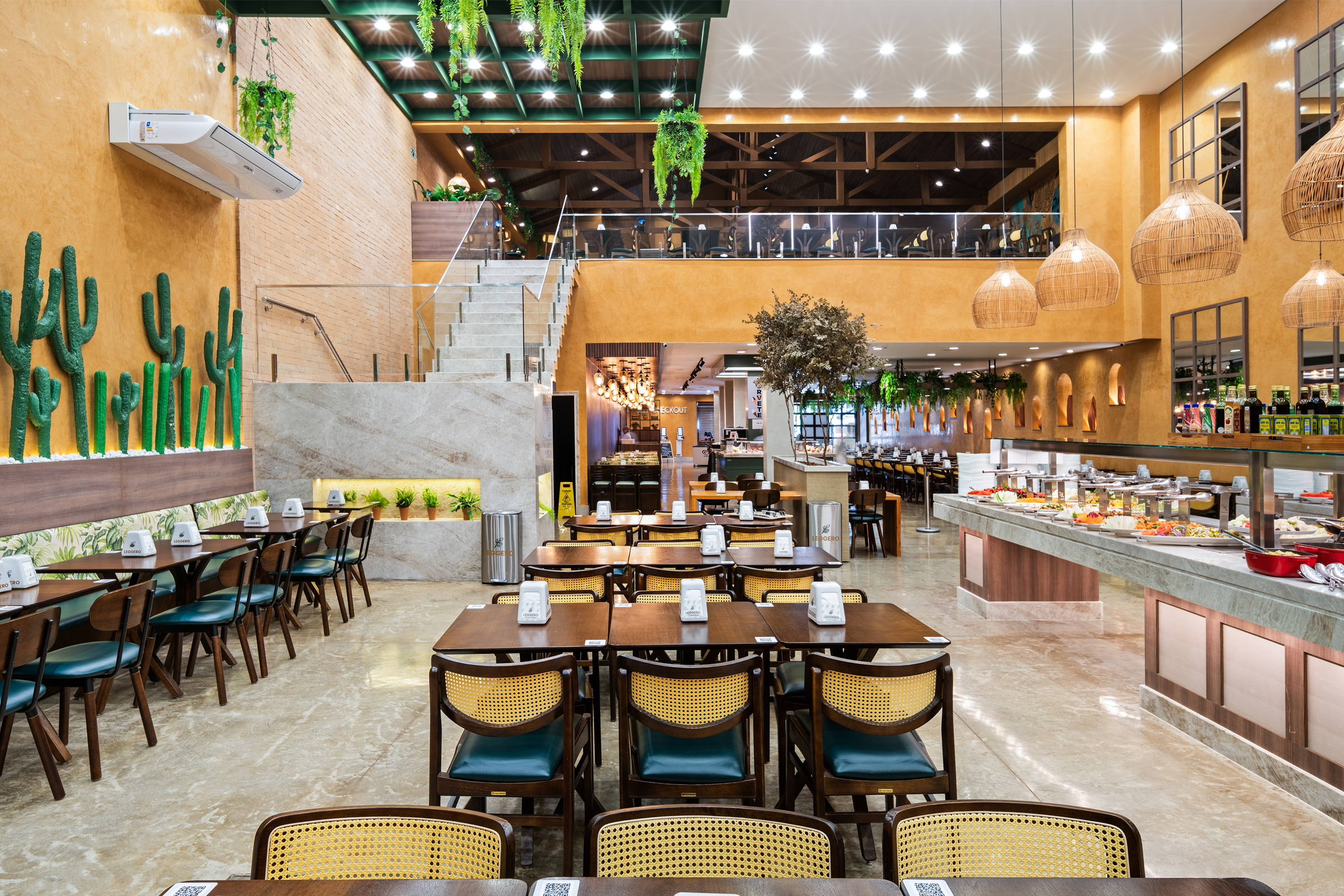 Vista geral do salão de um restaurante de buffet amplo e bem iluminado com decoração rústica e moderna. À direita, estende-se um longo balcão de buffet self-service com tampo de pedra clara, oferecendo diversas opções de pratos quentes e saladas em travessas coloridas; acima dele, há uma decoração inusitada com uma bicicleta verde suspensa. No centro e à esquerda, organizam-se várias mesas de madeira com cadeiras de encosto de palha e assentos escuros.