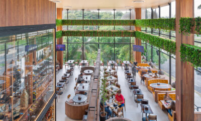 Vista aérea de um restaurante amplo com pé-direito duplo e arquitetura contemporânea. O salão principal possui piso claro e é organizado com diferentes tipos de mesas de madeira, incluindo booths circulares estofados em cinza no centro e sofás curvos de couro caramelo dispostos ao longo da lateral direita. Grandes pilares revestidos de madeira estruturam o espaço, decorado com folhagens verdes suspensas em vigas elevadas. Ao fundo e à direita, paredes de vidro integram o ambiente à vegetação externa, proporcionando iluminação natural.