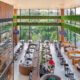 Vista aérea de um restaurante amplo com pé-direito duplo e arquitetura contemporânea. O salão principal possui piso claro e é organizado com diferentes tipos de mesas de madeira, incluindo booths circulares estofados em cinza no centro e sofás curvos de couro caramelo dispostos ao longo da lateral direita. Grandes pilares revestidos de madeira estruturam o espaço, decorado com folhagens verdes suspensas em vigas elevadas. Ao fundo e à direita, paredes de vidro integram o ambiente à vegetação externa, proporcionando iluminação natural.