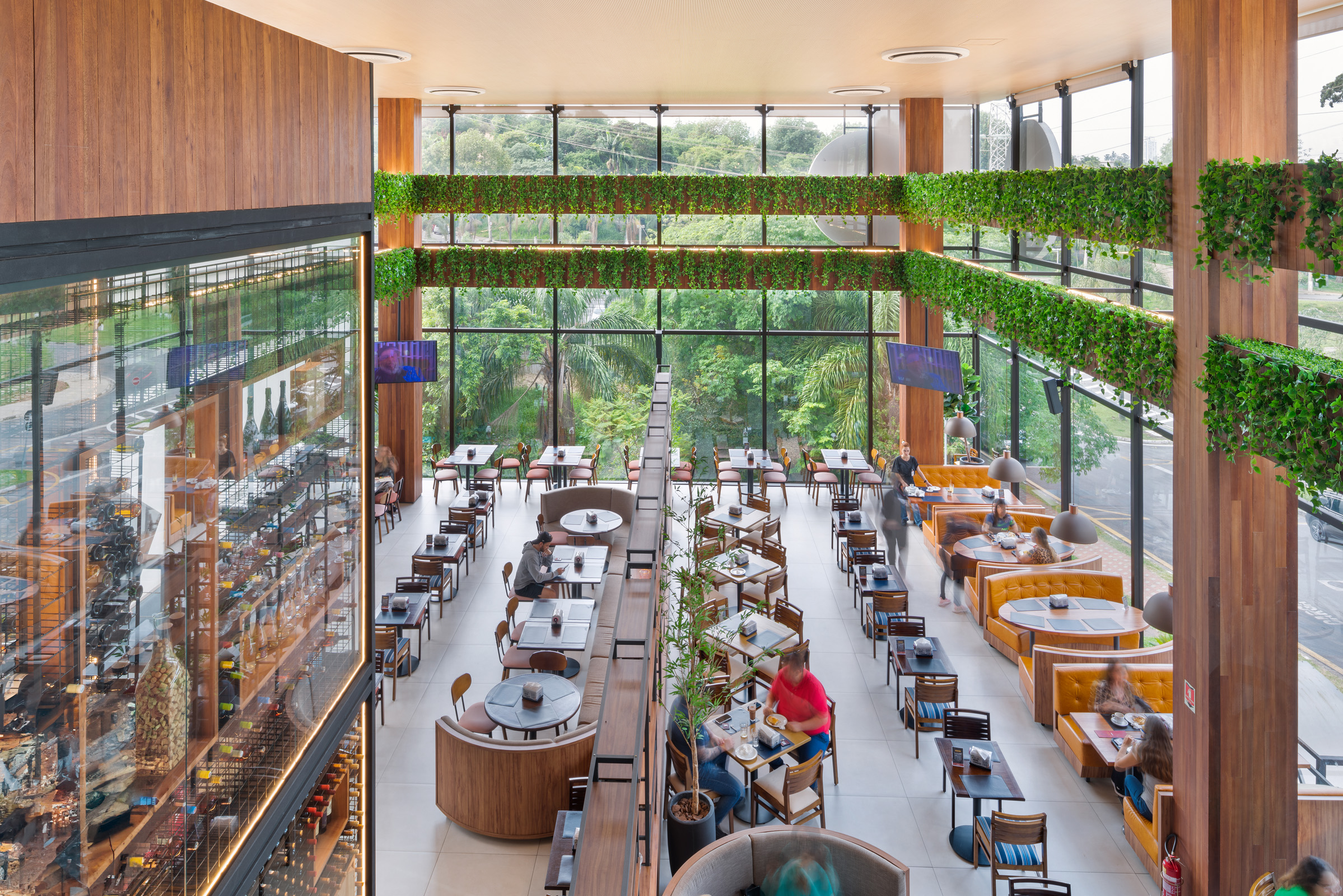 Vista aérea de um restaurante amplo com pé-direito duplo e arquitetura contemporânea. O salão principal possui piso claro e é organizado com diferentes tipos de mesas de madeira, incluindo booths circulares estofados em cinza no centro e sofás curvos de couro caramelo dispostos ao longo da lateral direita. Grandes pilares revestidos de madeira estruturam o espaço, decorado com folhagens verdes suspensas em vigas elevadas. Ao fundo e à direita, paredes de vidro integram o ambiente à vegetação externa, proporcionando iluminação natural.