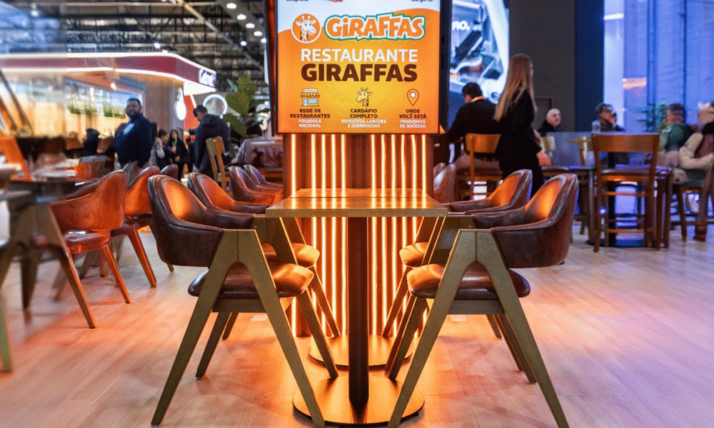 Área de refeições de um restaurante Giraffas com cadeiras de pele castanha e iluminação alaranjada sob um painel publicitário.