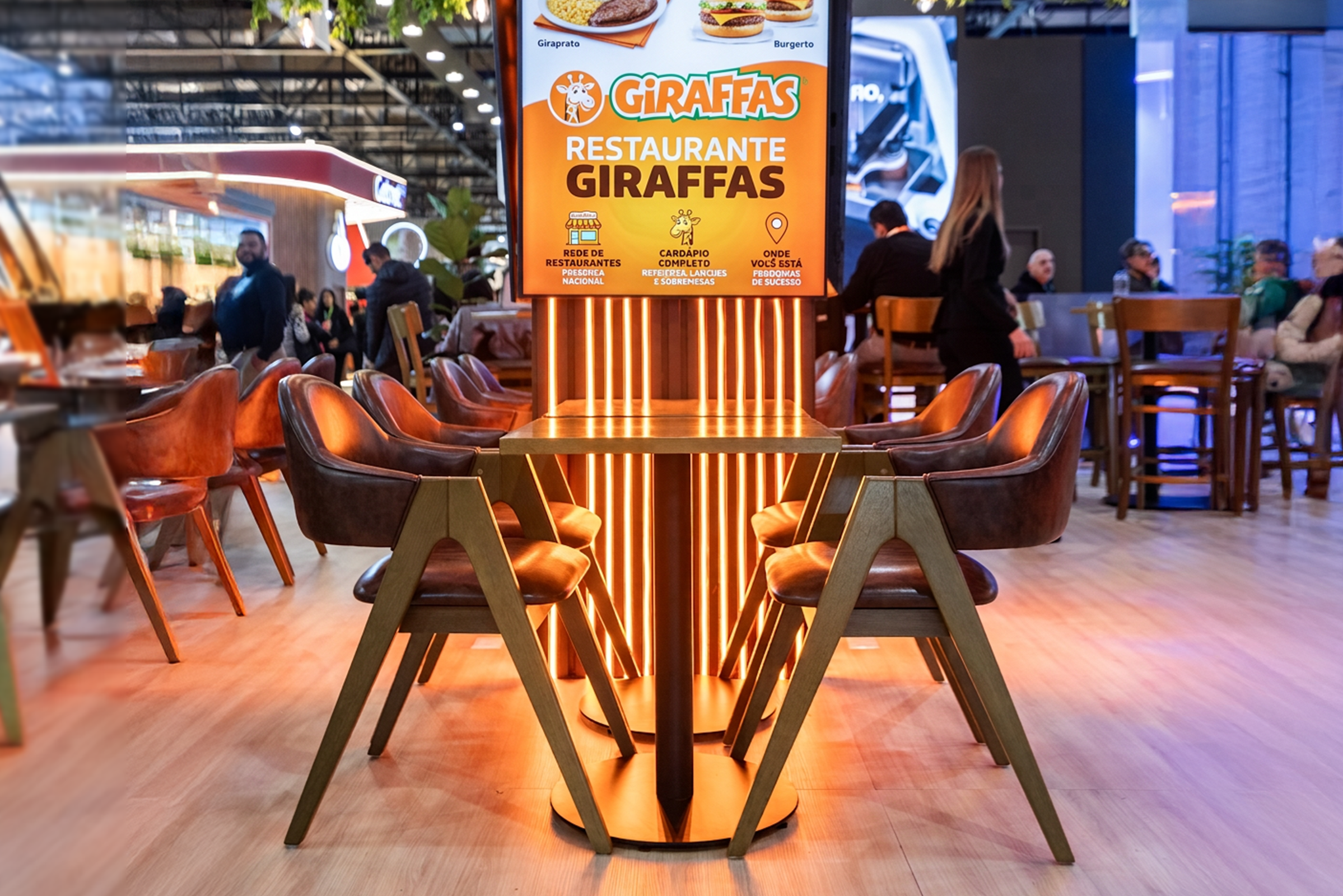 Área de refeições de um restaurante Giraffas com cadeiras de pele castanha e iluminação alaranjada sob um painel publicitário.