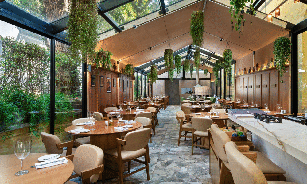 Interior de um restaurante moderno e luminoso, com teto de vidro, plantas suspensas, mesas redondas de madeira e um balcão em mármore.