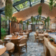 Interior de um restaurante moderno e luminoso, com teto de vidro, plantas suspensas, mesas redondas de madeira e um balcão em mármore.