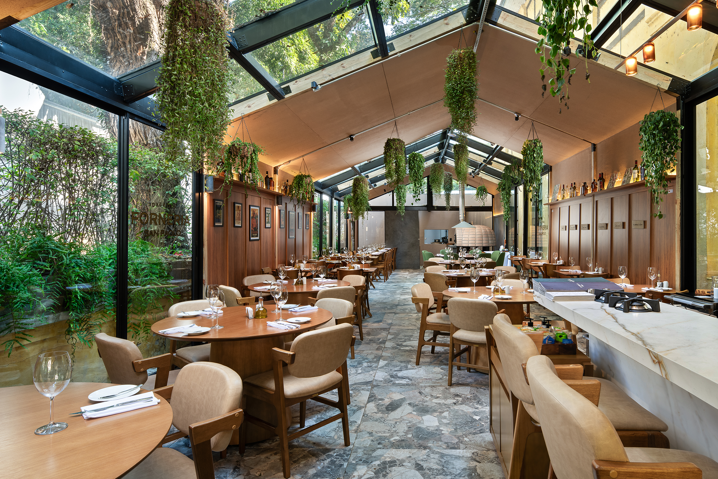 Interior de um restaurante moderno e luminoso, com teto de vidro, plantas suspensas, mesas redondas de madeira e um balcão em mármore.
