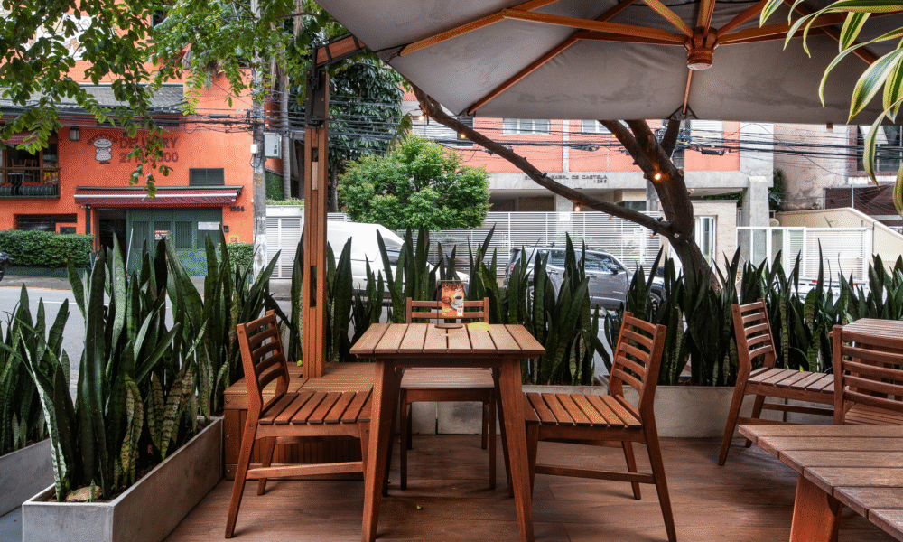 Esplanada ao ar livre com mesas e cadeiras de madeira sob um guarda-sol cinzento, rodeada por plantas em vasos e com vista para a rua.