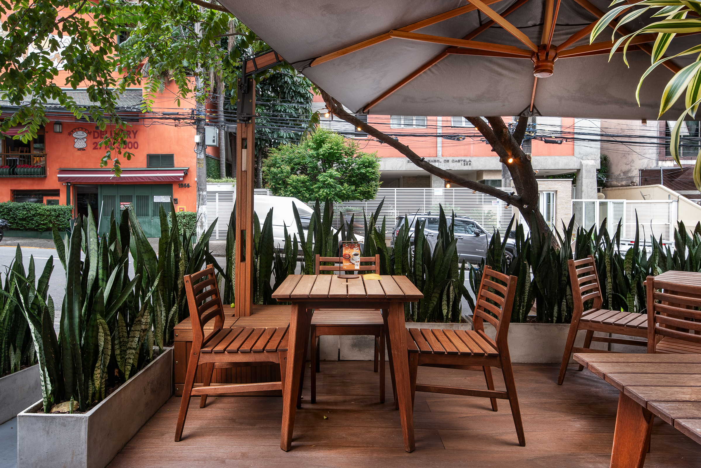 Esplanada ao ar livre com mesas e cadeiras de madeira sob um guarda-sol cinzento, rodeada por plantas em vasos e com vista para a rua.